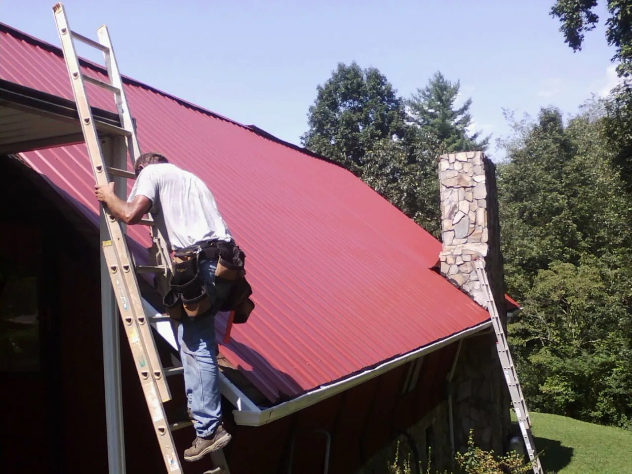 Roofer on a ladder beside a red metal roof and stone chimney — Soffit & Fascia Repair by Roofing Brothers in Paulden